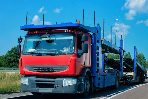 A red and blue car carrier truck, representing top-tier Car Transport Orangevale services, is parked on the side of a road under a blue sky with scattered clouds. Trees and greenery are visible in the background.