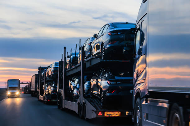 A car carrier truck loaded with new cars drives down a highway at sunset near Orangevale, CA, showcasing Car Transport Orangevale services, with other trucks visible in the background.